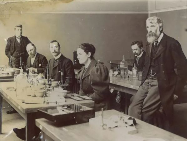 Men and women from the Victorian era stand around laboratory benches