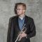 School of Music alumni Christopher Young. Christopher stands against a grey background and is looking to the left of the frame. He is holding his trumpet and wears a blue shirt and dark blazer.