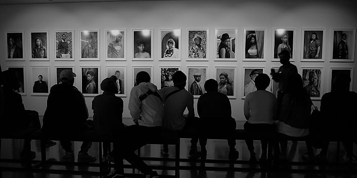 A black and white photo of people sitting on a bench from behind. In front of them are photographs.