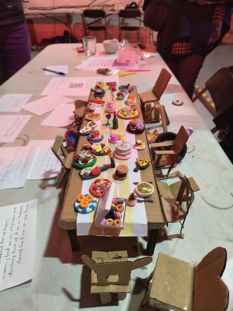 Miniature model of a dining table laden with food and chairs, set on a table and surrounded by written notes.