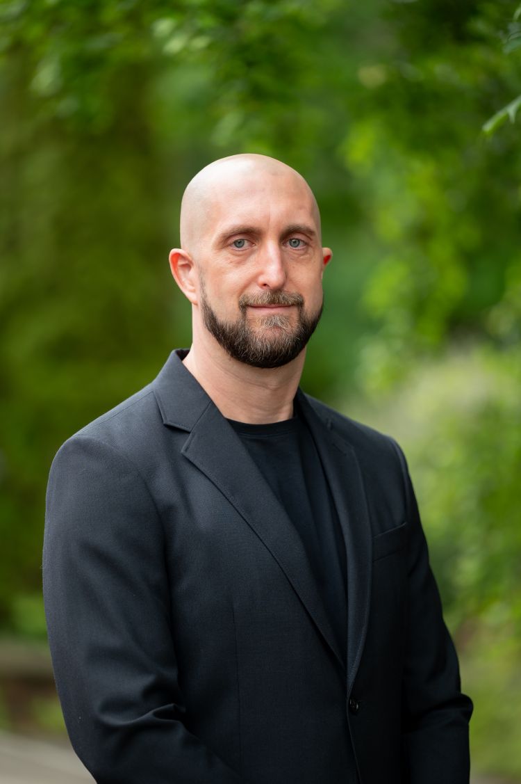 A man in a black suit smiles for the camera against a background of trees 