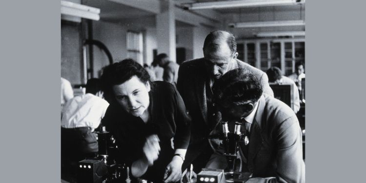 A black and white photograph showing a woman and two men bending over a microscope in a lab.