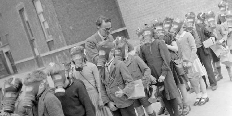 Image of school children wearing gas masks standing in a queue. A man, presumably their teacher, is looking over them.