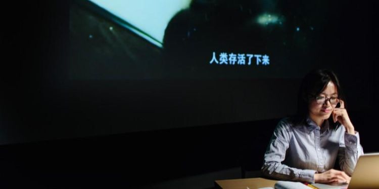 Student works thoughtfully at a desk beside a laptop as a dark film with Chinese subtitles is projected on the wall behind her
