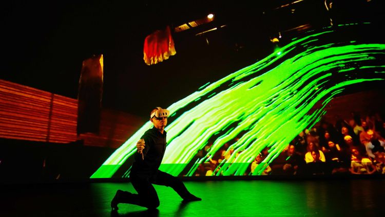 A dancing in a black suit kneeling on a stage with a VR headset.