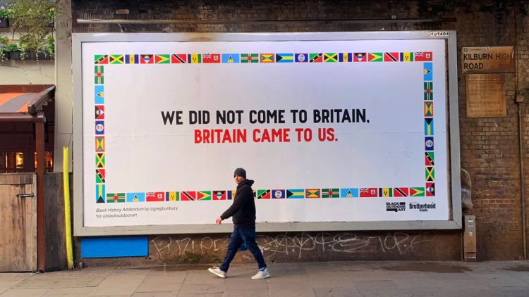 man walking in front of a billboard that states 'We did not come to Britain. Britain came to us.'