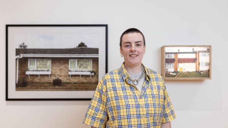 FAHACS student George Storm Fletcher photographed in front of their work featured at Leeds Art Gallery for Leeds Artists Show 2023. George is looking directly into the camera and smiling. To the left and right of George are two pieces of their work.