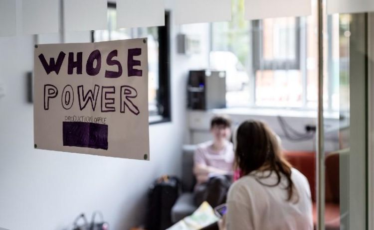 Two people in a room with a sign on the window that reads Whose Power production office.