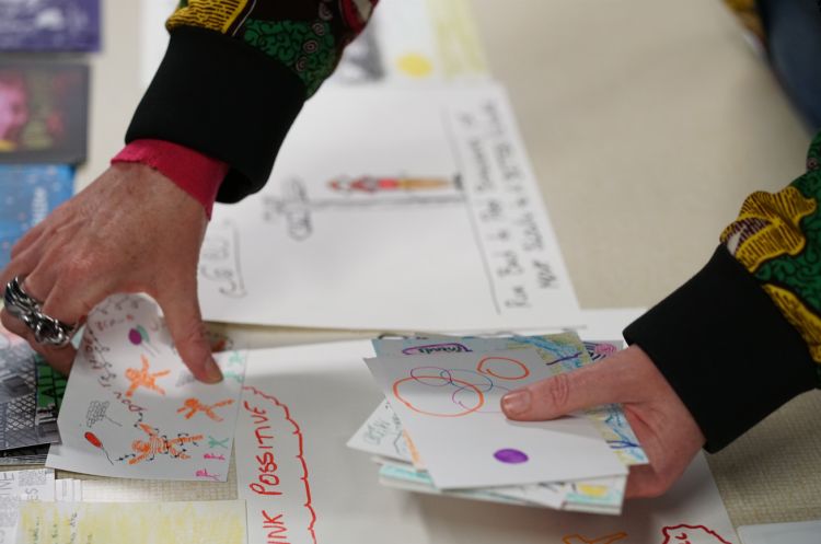 Close up of a woman’s hands holding a piece of paper featuring a detailed drawing. There are posters and drawings on the table beneath her hands.