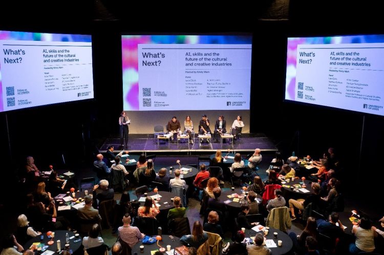 Six people sit on a stage in front of an audience assembled around tables