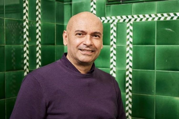 Cesar stands in front of a green tiled background, wearing a purple jumper, in the School of Fine Art, History of Art and Cultural Studies.