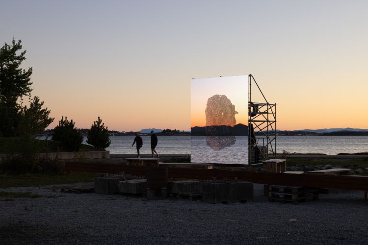 It's dusk, 2 people are walking by a wide river. There is a screen (held up by scaffolding) with a projection of what looks like a large boulder. You can see through the projection to the river beyond.