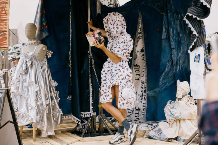 A person dressed in a all in one hooded dress (white with a pattern) leaning against a
 pole which is holding up lots of fabric in the shape of a tent. A mannequin is standing to the left dressed in a white silk dress with frills.