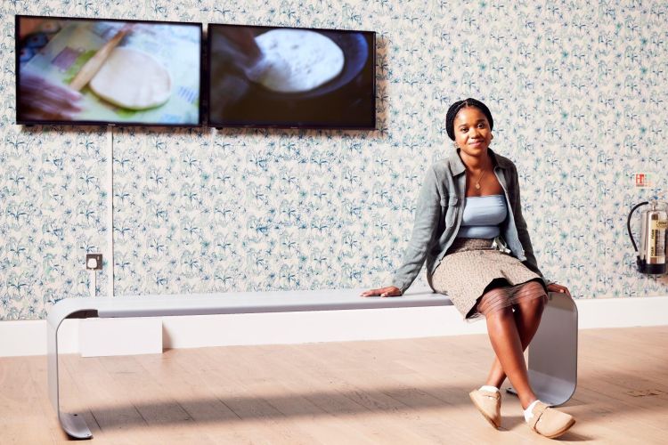 Artist Kalisha Piper-Cheddie seated in a gallery with her video installation and wallpaper