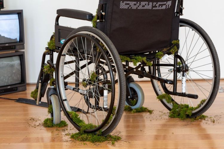 a black nhs wheelchair is in the foreground, behind it a stack of six crt monitors are playing a film. Moss appears to be growing over the wheelchair, around the tyres and belting. The back panel is embroidered with three rows of white cross stitch