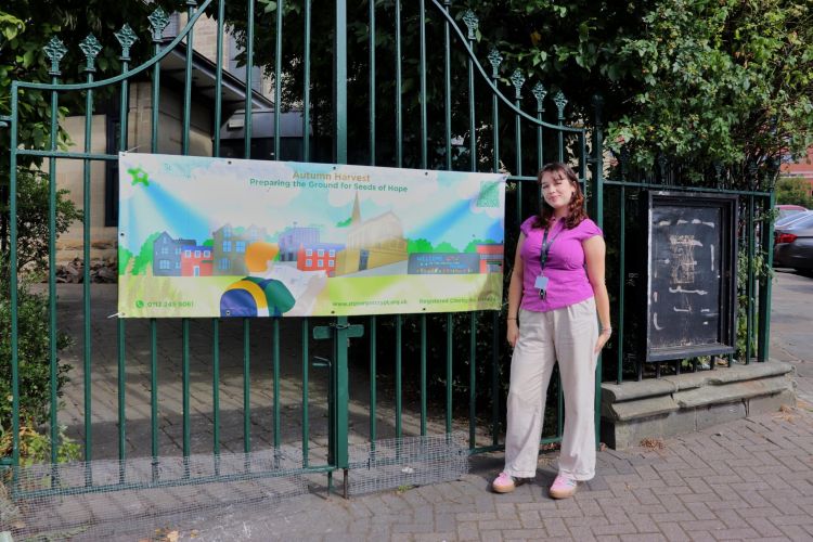 Madalena Teague stands outside gates with a banner advertising the Autumn Harvest