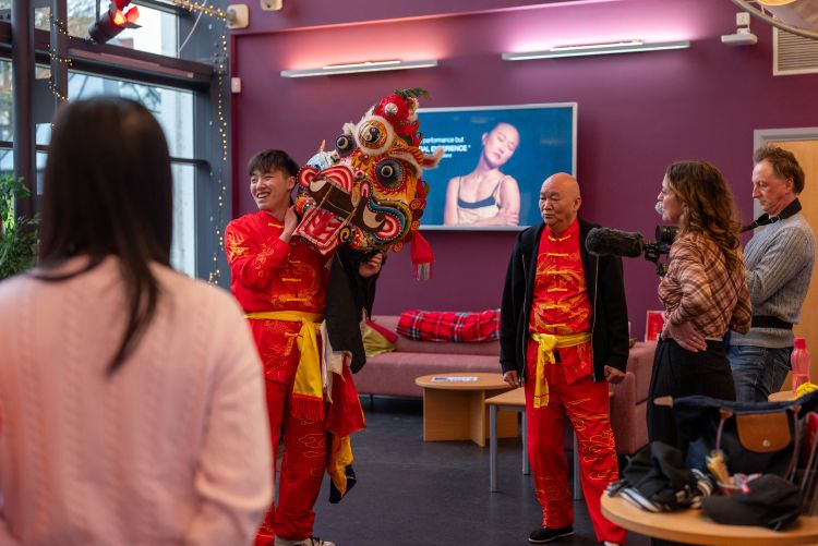 2 Chinese men dressed in traditional performing arts costumes holding a Chinese dragon puppet in a theatre foyer