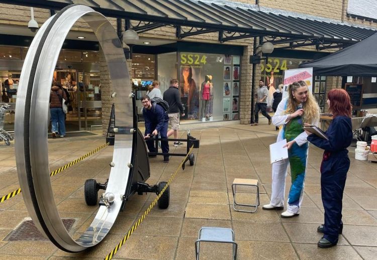 Sculpture of a wheel in a town centre street on the left with two people talking to each other on the right of the photo