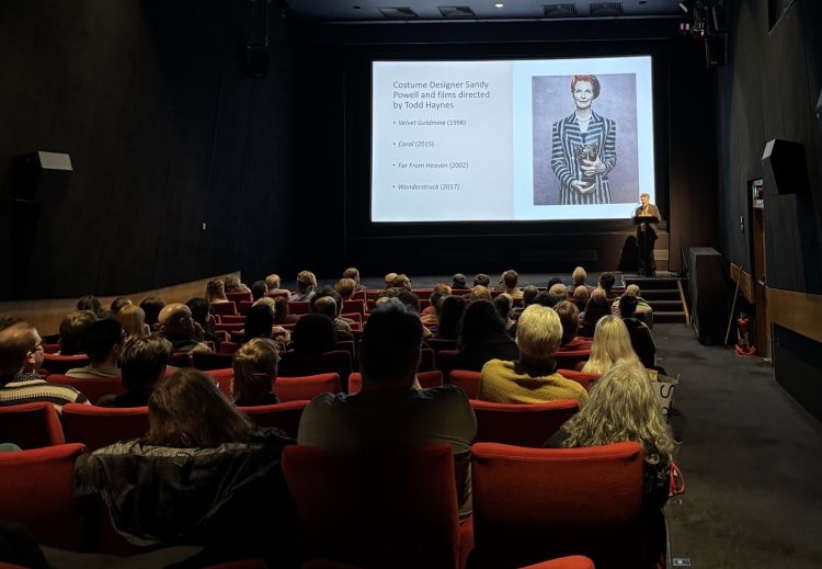 Standing at a lectern on a stage, a woman delivers a speech to an audience in a cinema 