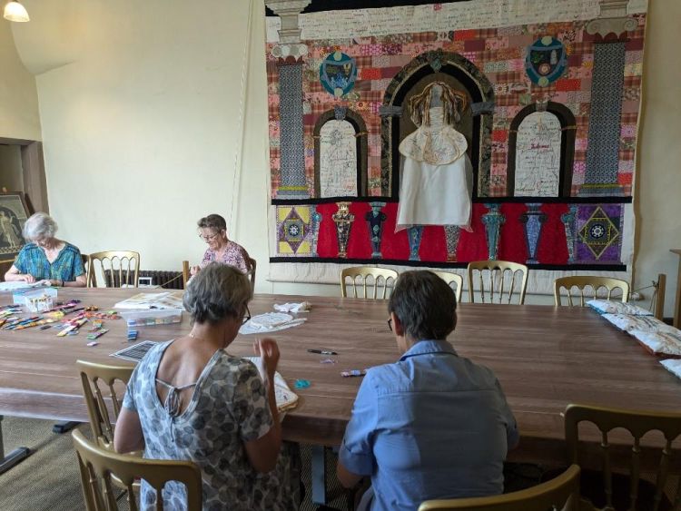 People at a table doing embroidery with large tapestry in the background