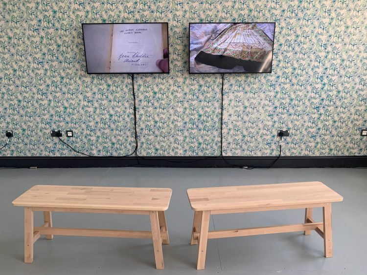 Artwork by Kalisha Piper-Cheddie: two video monitors on a wall covered in wallpaper with two benches in foreground