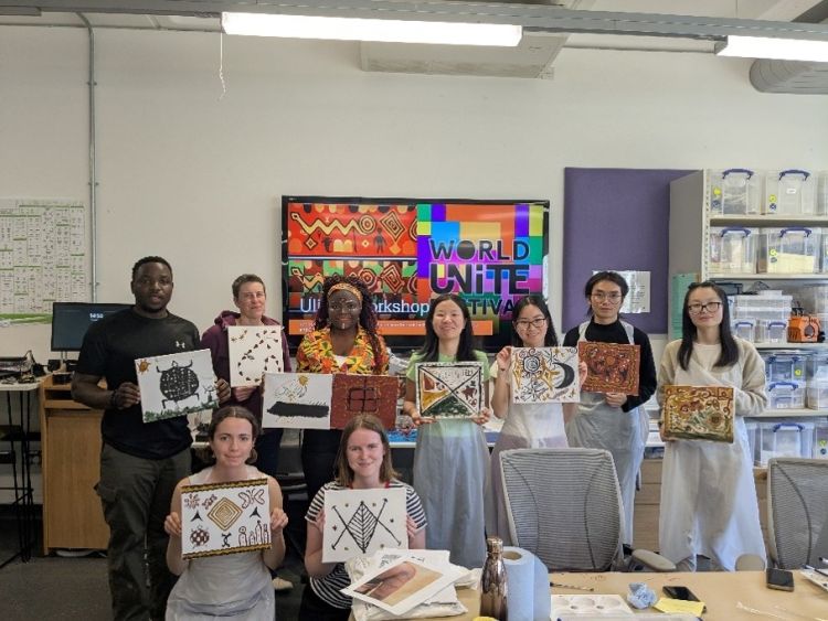 A group of people in a classroom smile for the camera, holding up artworks from a workshop they have taken part in