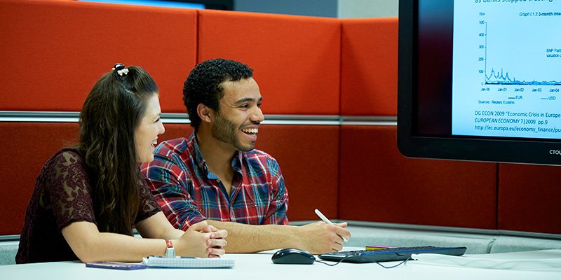 Two students working at a booth in the library with big screen presentation technology.