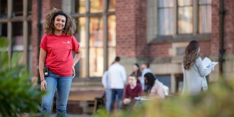 A student ambassador wearing red t shirt and blue jeans stood in front of a red brick building with students blurred out in the background and blurred greenery in the foreground