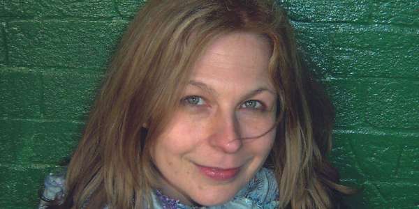 Jill Crossland vocal and instrumental teacher in the School of Music. Jill is looking directly at the camera and smiling, she stands in front of a brick wall that has been painted green.
