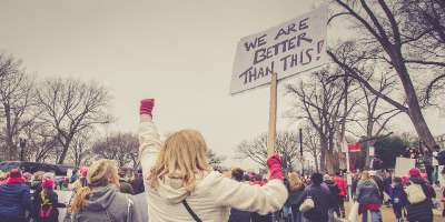 Group of protestors with the back of a woman in foreground holding up a sign and other hand in a fist in the air