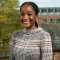Student Shanté Thompson. Shanté stands in front of an out of focus background, to the right is a university building and to the left is a tree. Shanté is smiling and wears a neutral check high neck top.