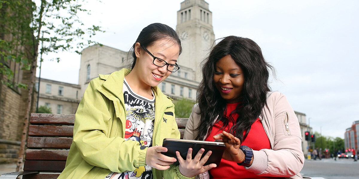 Two students sat on a bench reading from a tablet device