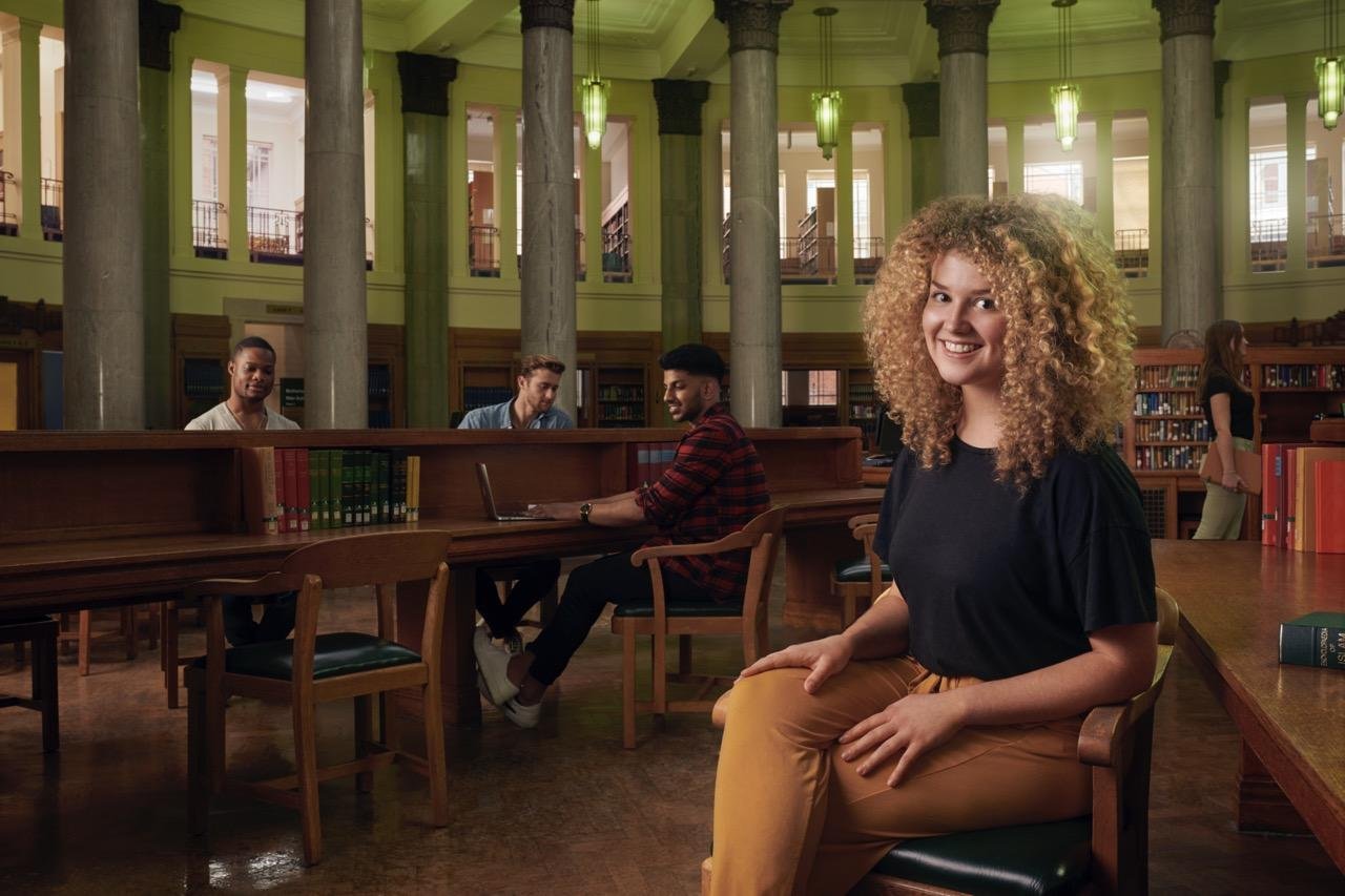 A female student sits in the Brotherton library facing the camera with a smile. In the background other students can be seen working.