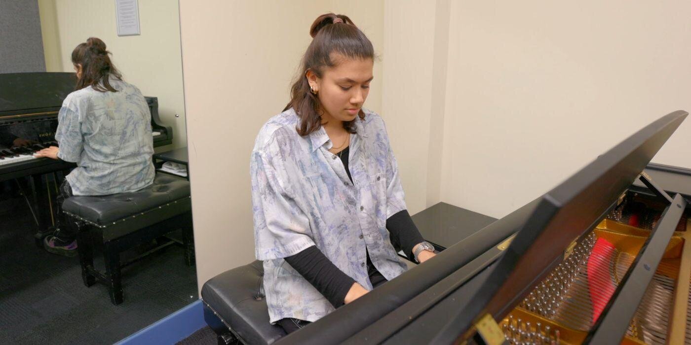 School of Music rep, Georgie, who is sat at a piano in one of the practice rooms rehearsing a piece of music. Georgie is looking down at the piano.