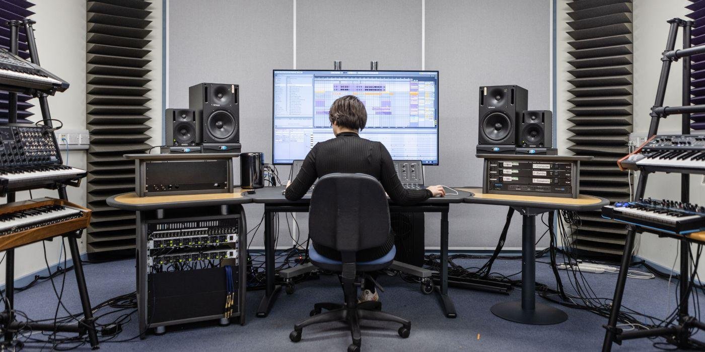 Student using one of the electronic studios in the School of Music. They are using software on a screen and are surrounded by keyboards, speakers, and other electronic equipment. They have their back to the camera.