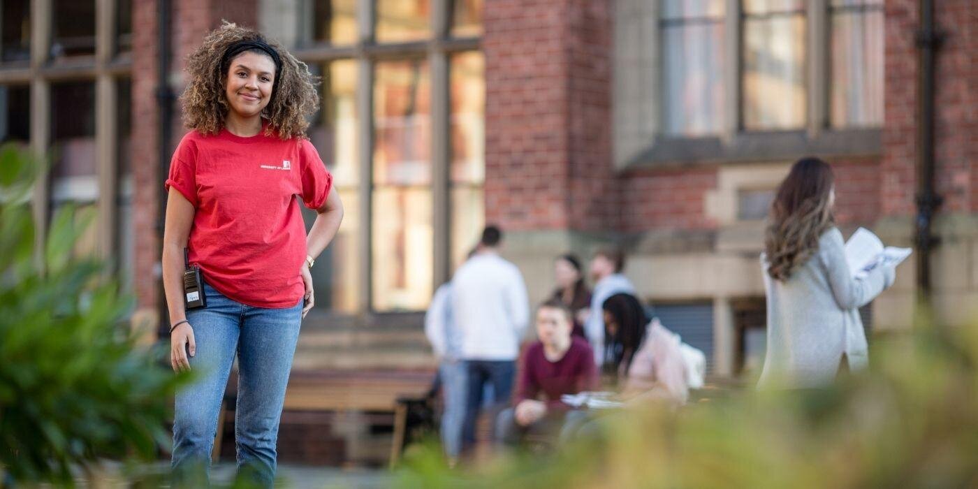 A student ambassador wearing red t shirt and blue jeans stood in front of a red brick building with students blurred out in the background and blurred greenery in the foreground