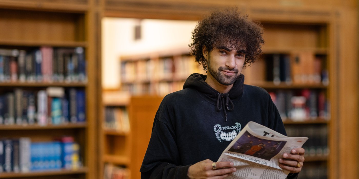A male student smiles to the camera, in the background are the bookcases of the Brotherton Library.