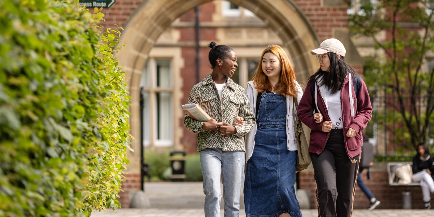 Three female students walk through campus arm in arm, they are laughing and talking. In the background is the redbrick arch of Clothworker's Court.