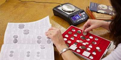 Person sorting ancient coins into a coin presentation box using a reference book and scales.