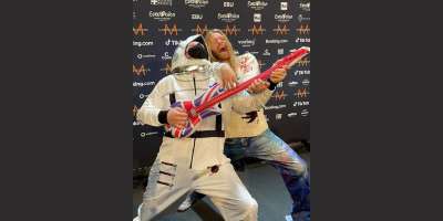 Sam Ryder in front of a backdrop with Eurovision logos on it. He's with somebody dressed as a spaceman with a blow up guitar.