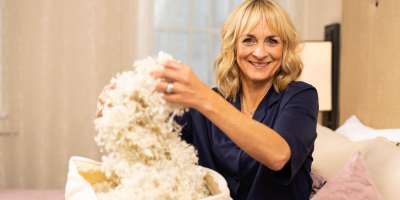 Former BBC presenter Louise Minchin sitting on a bed, smiling, holding a bag of wool
Picture via Woolroom