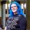 Lizzie is stood in the Brotherton library. She has bright blue hair, and is wearing glasses and a black jumper with a floral pattern. She is smiling at the camera.
