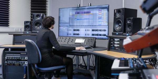 Student using one of the electronic studios in the School of Music. She is sat at a large screen using software and is surrounded by keyboards, speakers and other equipment.