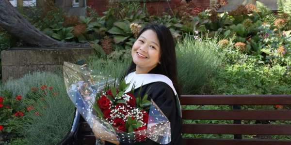 Novasari sitting on a bench, dressed in graduation attire holding flowers and smiling.