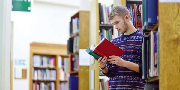 A male student leans against a library bookcase, an open book in his hands as he reads.