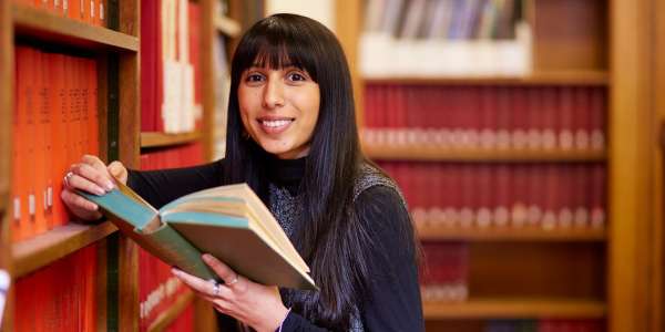 A female student leans against a bookcase, a book open in her hands, as she smiles at the camera.